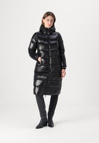 Gevoerde zwarte jas met een glanzende afwerking, knielang, hoge kraag, ritsluiting en zijzakken. Draag hem over donkere jeans en enkellaarsjes.
