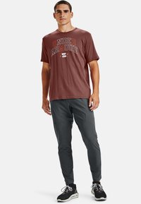 Maroons krāsas Under Armour T-krekls ar baltu un oranžu logo, kombinēts ar tumši pelēkam šauriem biksēm. Modelis valkā melnus un baltus sporta apavus.