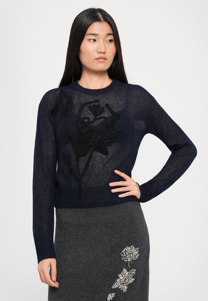 Una donna con lunghi capelli scuri indossa un maglione trasparente blu navy con ricami floreali neri e una gonna grigia con decorazioni floreali bianche.