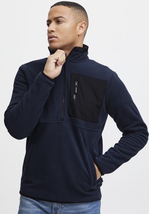 Marineblauer Fleece-Pullover mit halbem Reißverschluss, ausgestattet mit einer schwarzen Reißverschlusstasche auf der Brust, seitlichen Handtaschen und gerippten Bündchen. Glatte Textur.