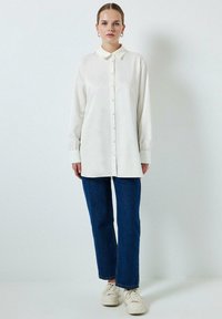 Camisa blanca de manga larga con cierre de botones y cuello, combinada con jeans rectos azules y zapatillas blancas. Textura suave y corte relajado.