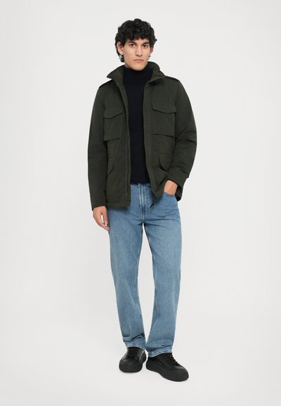 Dunkelgrüne Jacke mit Kragen und vier Vorderaschen, getragen über einem schwarzen Rollkragenpullover, kombiniert mit hellblauen Jeans und schwarzen Schuhen.
