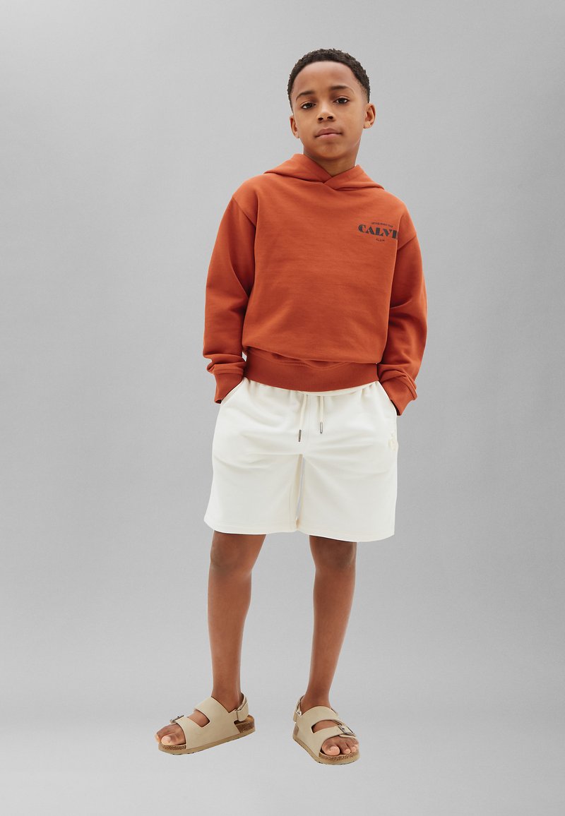 Garçon debout, les mains dans les poches, portant un sweat à capuche couleur rouille, un short blanc et des sandales beiges sur fond gris.