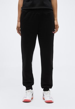 Person iført sorte træningsbukser med pink logo på låret og hvide sportssko med pink såler, stående mod en ensfarvet baggrund.