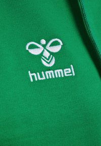 Zielony materiał o gładkiej fakturze zdobiony jest białym haftowanym logo w kształcie pszczoły oraz słowem "hummel" poniżej.