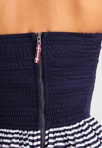 Dunkelblaues Kleid mit gerafftem Oberteil und dunkelblau/weiß gestreiftem Rock. Verfügt über einen sichtbaren Reißverschluss mit einem farbigen Zipper. Die Textur wirkt glatt.