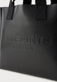 Saco tote de pele preta com o logótipo em relevo "ALLSAINTS LONDON", apresentando alças de tecido preto robustas e um design elegante e minimalista.