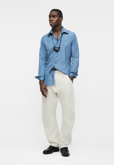 Άνδρας που φοράει μπλε ριγέ πουκάμισο με κουμπιά, λευκό φαρδύ παντελόνι, μαύρα παπούτσια και μικρό μαύρο κρεμαστό τσαντάκι, στέκεται με το ένα χέρι στην τσέπη.