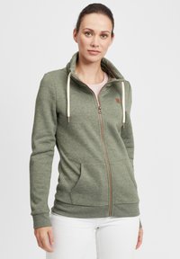 Grønn zip-up hettegenser laget av mykt materiale, med høy krage, kremfargede snorer, to frontlommer og en subtil logo-detalj.