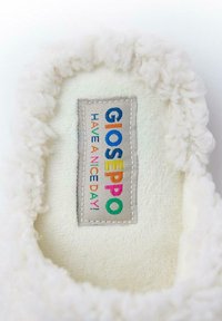 Interno di una pantofola bianca e soffice con una consistenza morbida; presenta un'etichetta colorata con la scritta "GIOSEPPO HAVE A NICE DAY!" su uno sfondo grigio chiaro.
