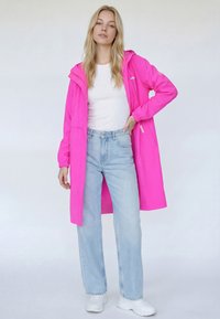 Pardesiu lung roz cu glugă, asortat cu un top alb și blugi bleu deschis. Jacheta are manșete elastice și buzunare laterale.