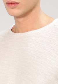 Jasno beżowy sweter o fakturze z okrągłym dekoltem, z delikatnym wzorem tkanym i gładką powierzchnią. Widoczne są krótkie rękawy.