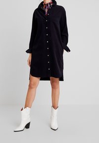Abito camicia in corda di colore blu navy scuro, con bottoni, due tasche sul petto, maniche lunghe e spacchi laterali, abbinato a stivali da cowboy bianchi a punta.