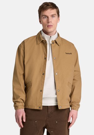 Mand iført en beige Timberland jakke over en hvid kvartzip pullover og mørkebrune bukser, stående mod en hvid baggrund.