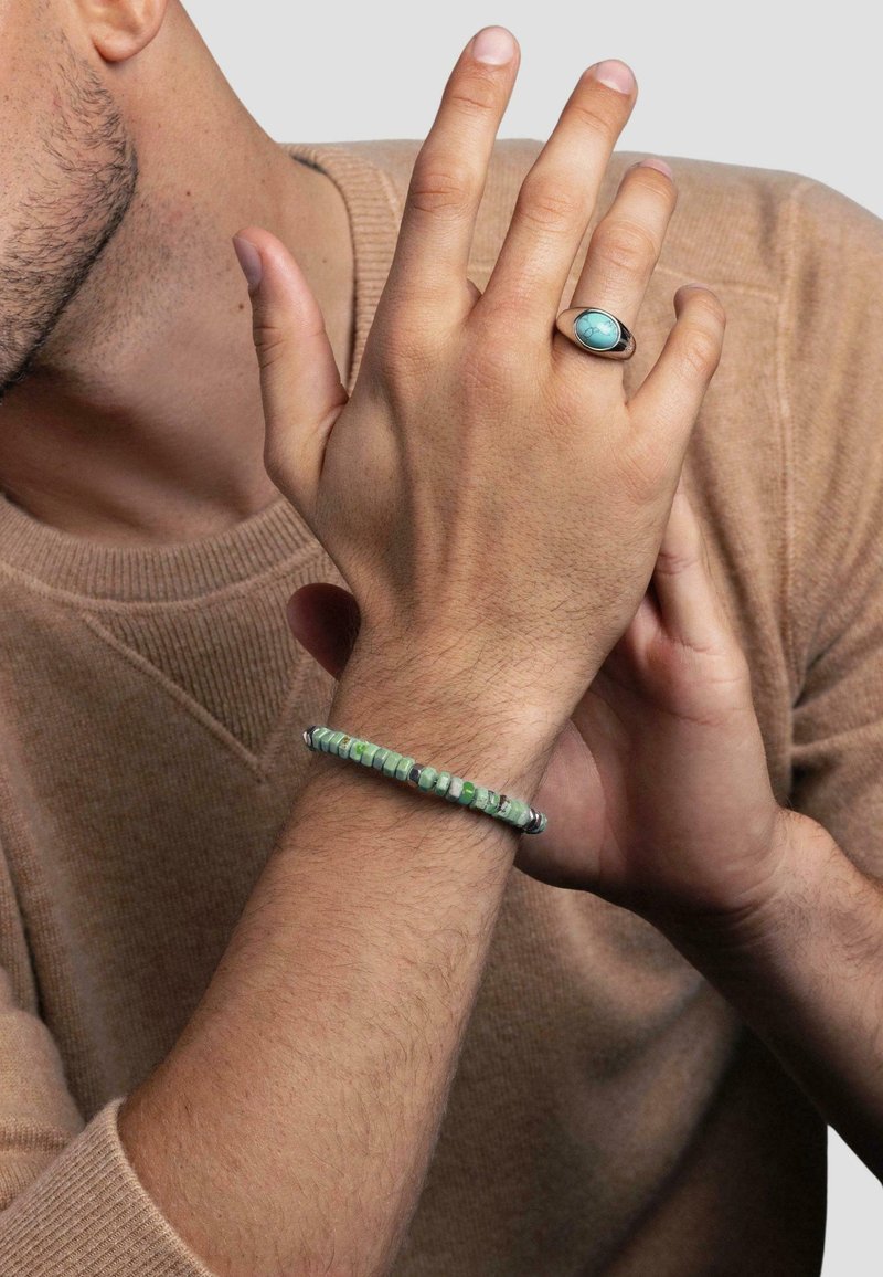 Beaded bracelet with green and black patterned stones, worn on the wrist. Silver-tone ring with an oval turquoise stone on the hand.