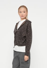 Sweatshirt cropped com fecho em padrão de leopardos, tecido cinza escuro com detalhes castanhos, apresentando um bolso frontal e um corte descontraído, combinado com uma T-shirt branca.