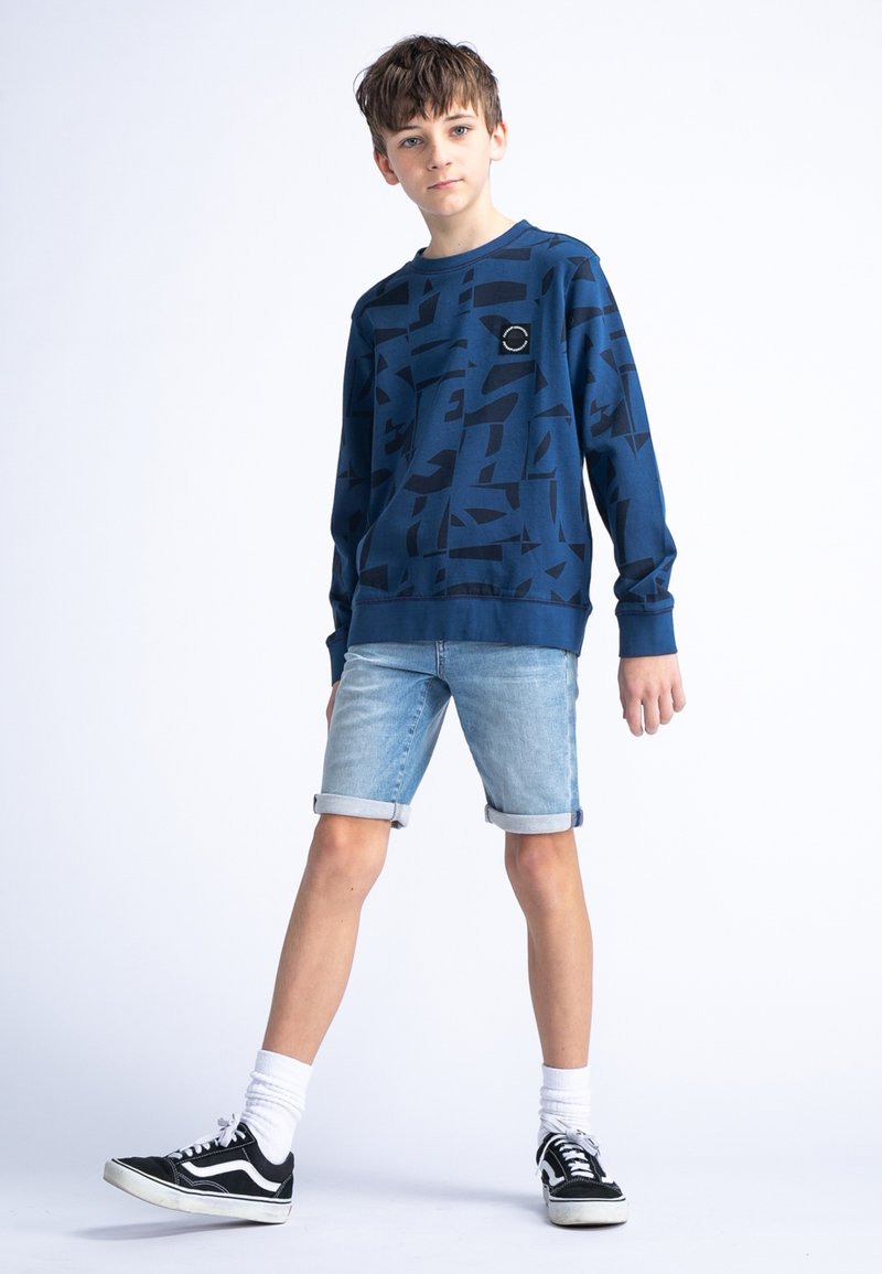 Felpa blu navy con motivo astratto nero, colletto a girocollo, abbinata a pantaloni shorts in denim chiaro e scarpe da ginnastica nere. Calzini bianchi visibili.