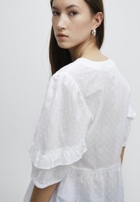 Blouse blanche avec des pois texturés, des détails en volants sur les manches et une coupe décontractée. Tissu doux et léger avec un col rond.