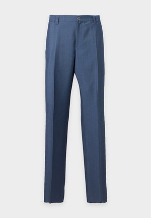 Jonge man staat met één hand in zijn zak, draagt een marineblauwe polo met korte mouwen, blauwe broek en zwarte schoenen met gouden accenten.