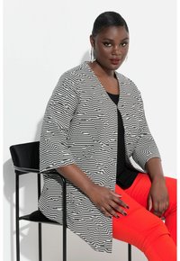 Cardigan a righe bianche e nere con texture floreale, con apertura frontale, maniche a tre quarti, indossato su una maglia nera e pantaloni rossi. Seduto su una sedia.