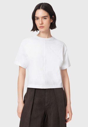 Femme aux cheveux courts et foncés portant un crop top blanc uni à manches courtes et un pantalon marron foncé taille haute, debout devant un fond uni.