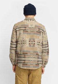 Beige gemustertes Hemd mit geometrischen Designs in Grau, Orange und Schwarz; lange Ärmel und abgerundeter Saum; kombiniert mit khakifarbenen Hosen und einer marineblauen Mütze.