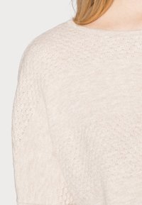 Pull en tricot beige clair avec un motif texturé et un col rond, fabriqué dans un matériau doux et léger.