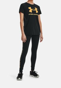 Persoon draagt een zwart Under Armour T-shirt met logo en zwarte legging met verticale Under Armour-tekst, gecombineerd met zwart-witte sneakers.
