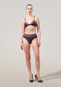 Kvinna står mot en neutral bakgrund iklädd genomskinlig lila lingerie och svarta högklackade skor, med blicken rakt fram och ett neutralt uttryck.