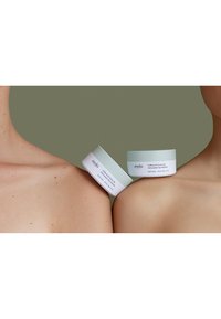 Due barattoli di crema per gli occhi sono esposti su uno sfondo verde tenue. I barattoli sono rotondi, di un verde chiaro con etichette bianche che riportano i dettagli del prodotto.