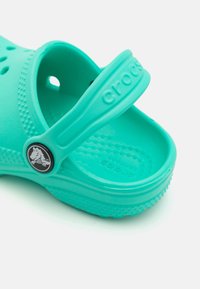 Turkio spalvos Crocs klumpės su kulkšnies dirželiu, perforuotu dizainu ir tekstūruota padu. Ant dirželio yra apvalus logotipas. Pagaminta iš lengvos medžiagos.
