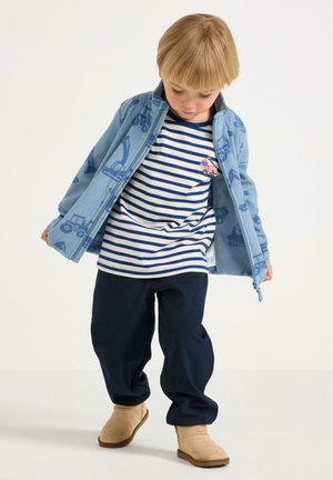 Bambino piccolo che guarda verso il basso, indossa una maglietta a righe blu, una giacca con zip azzurra chiaro con fantasie di veicoli, pantaloni scuri e stivali beige su sfondo bianco.