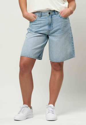 Henkilö, joka pukeutunut vaaleansinisiiin farkkushortseihin, valkoisiin lenkkareihin ja valkoiseen paitaan, kädet taskuissa, yksinkertaisen taustan edessä.