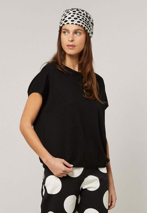 Vrouw draagt een zwart T-shirt met korte mouwen, zwarte broek met grote witte stippen, en een bijpassend wit hoofddoek met zwarte stippen.