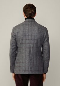 Blazer a quadri grigi con un design aderente, caratterizzato da un colletto scuro, cuciture sartoriali e una texture liscia. Bottoni minimalisti sui polsini.