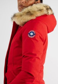 Parka rouge avec capuche en fourrure, dotée d'un patch logo rond sur la manche et d'un design ajusté. Extérieur lisse, chaud et isolé.