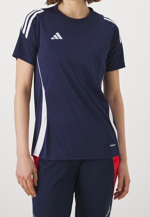 Personne portant un maillot de sport Adidas bleu marine avec des rayures blanches sur les épaules et les côtés, assorti à un short de sport bleu marine et rouge.