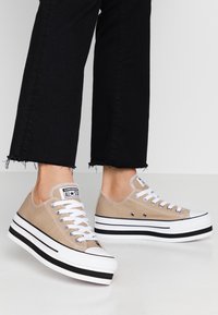 Nohy obuté v béžových platformových teniskách Converse All Star s bílými tkaničkami a černými pruhovanými podešvemi, doplněné o černé kalhoty s roztrhaným lemem.