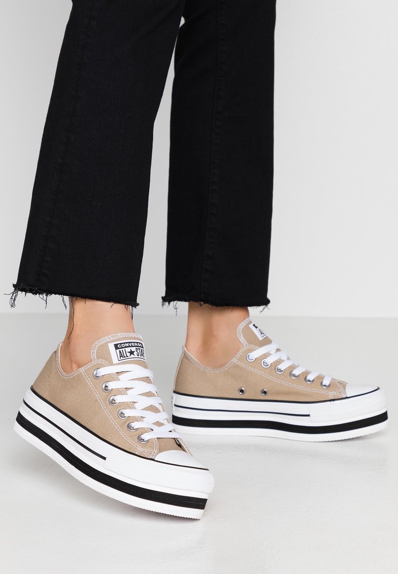 Nohy obuté v béžových platformových teniskách Converse All Star s bílými tkaničkami a černými pruhovanými podešvemi, doplněné o černé kalhoty s roztrhaným lemem.