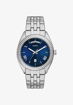 Montre Fossil argentée avec cadran bleu, chiffres romains, affichage du jour et de la date, bracelet en métal à maillons et lunette texturée.