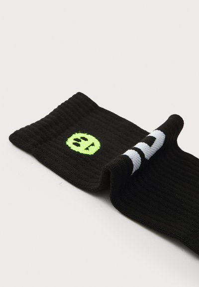 Chaussette noire côtelée avec un bord roulé, ornée d'un sourire circulaire néon vert et d'une inscription blanche sur le dessus. Tissu texturé.