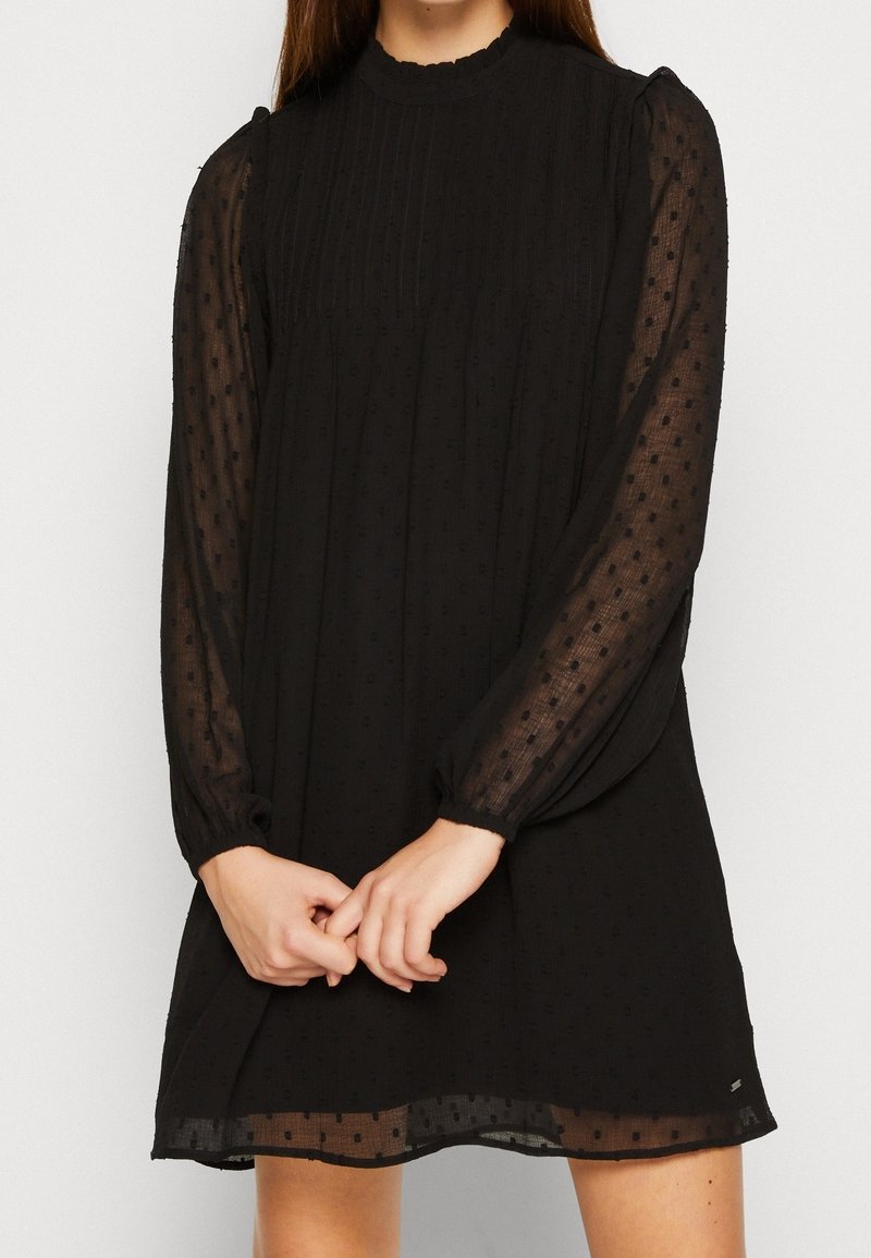 Femme portant une robe noire à manches longues avec des manches transparentes à pois et un col montant, mains jointes devant.