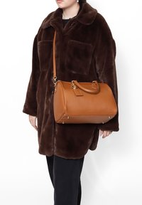 Cappotto in pelliccia sintetica marrone con grandi tasche, abbinato a una borsa in pelle beige con texture liscia e dettagli in oro, su un outfit nero.