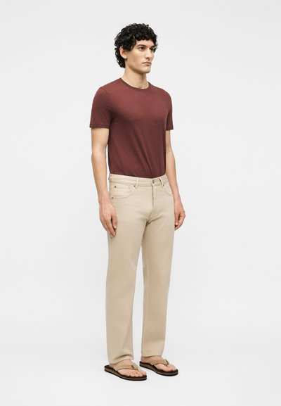 Homme aux cheveux bouclés portant une chemise marron à manches courtes, un pantalon beige et des tongs beiges, debout devant un fond blanc uni.