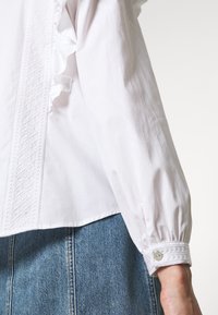 Blouse blanche à manches longues avec panneau vertical brodé et détail à volants, associée à une jupe en denim bleu.