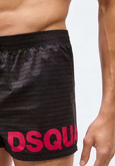 Gros plan sur une personne portant un short noir avec une ceinture élastique, affichant le texte répété "DSQUARED2" et de grandes lettres rouges "DSQUA".