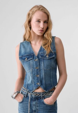 Blonde Frau mit welligem Haar trägt ärmellose Jeansweste und Jeans mit silbernem Kettengürtel, Hände in den Taschen, vor einfarbigem Hintergrund.