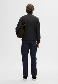 Donker sweatshirt met hoge kraag, gecombineerd met marineblauwe broek en zwarte sneakers. Een bruine tas is over de schouder geslingerd.