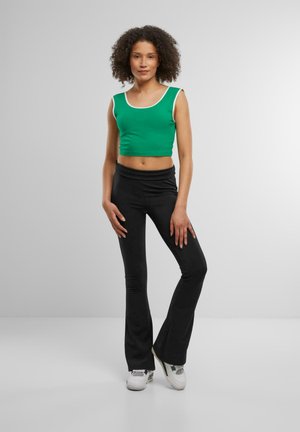 Top corto verde con rifiniture bianche, abbinato a pantaloni neri a zampa d'elefante. Modella che indossa sneakers bianche con accenti verdi sottili. Texture morbide.