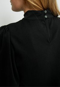 Top noir à col montant, avec des manches bouffantes et une fermeture à boutons dans le dos. Tissu doux avec une texture lisse et peu de détails de couture.